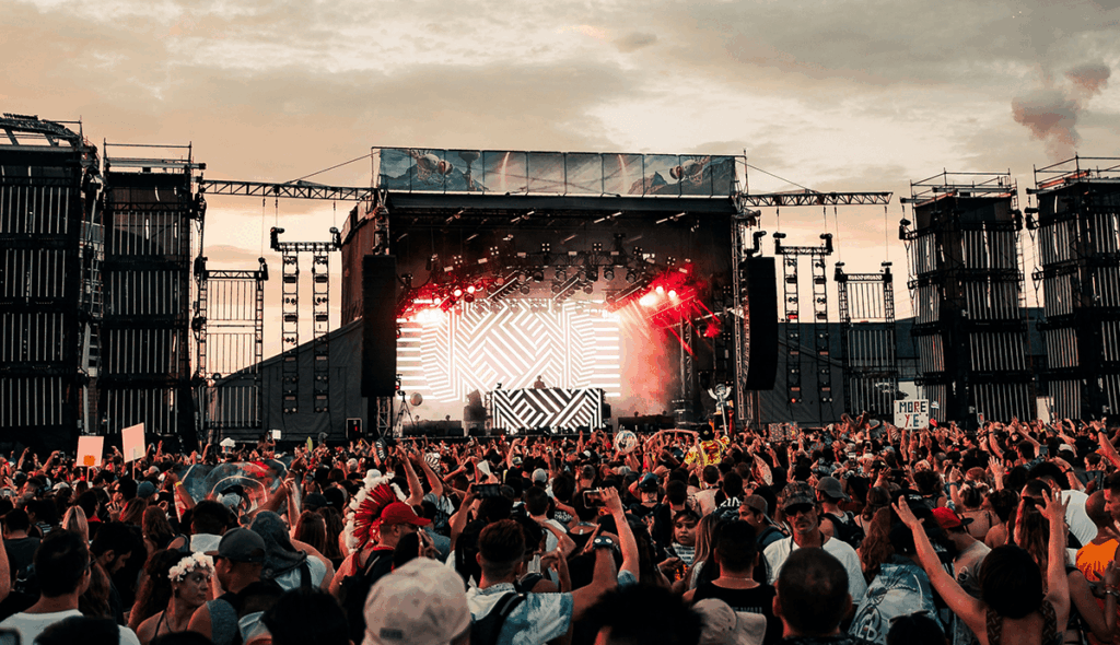 Gran escenario de festival de música al aire libre con pantalla LED de diseño geométrico en blanco y negro, luces rojas y humo escénico, rodeado de una multitud numerosa con los brazos en alto al atardecer.