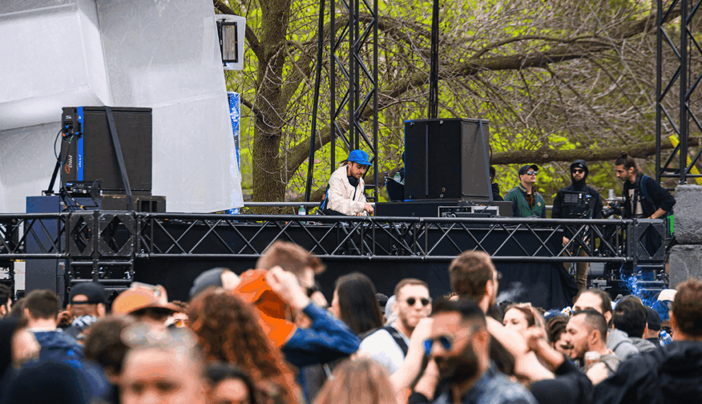 DJ con gorra azul actuando en un escenario al aire libre rodeado de altavoces profesionales y árboles en primavera, ante una audiencia numerosa disfrutando del evento durante el día.